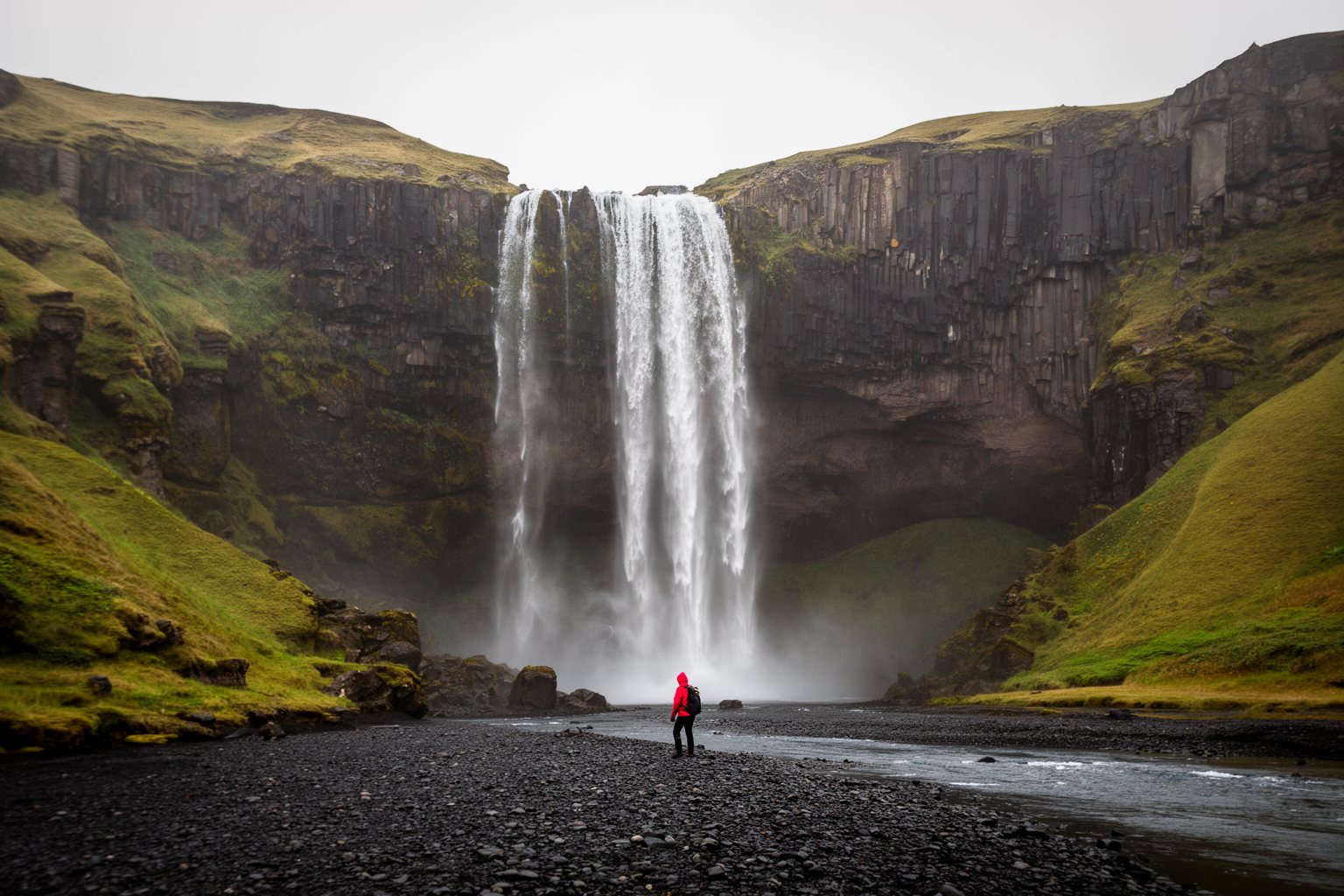 Hiker in a red jacket beneath a tall waterfall in a mossy Icelandic landscape