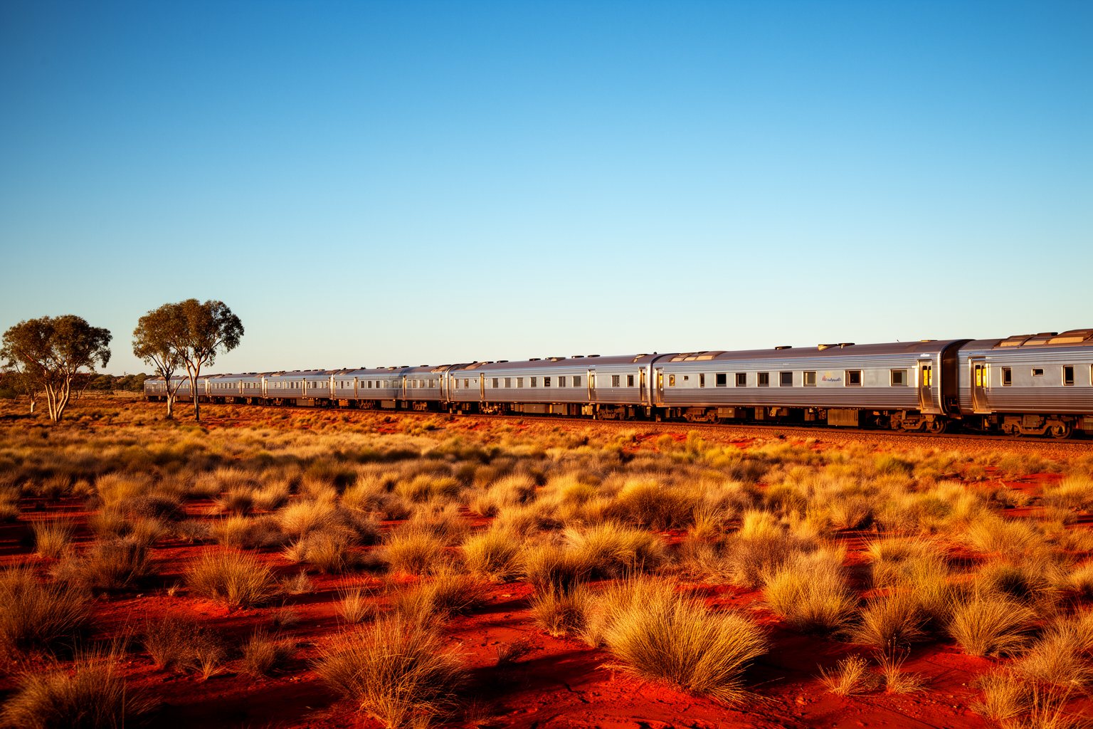 Riding the Rails: Unforgettable Train Journeys Around the World Long passenger train crossing the red Australian outback under a vast sky