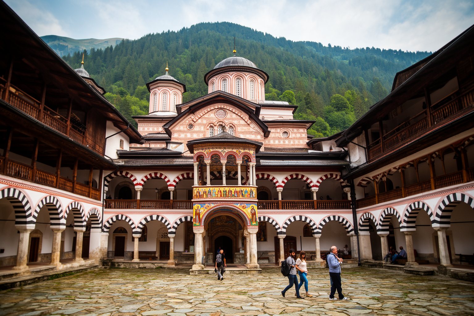 10 Unmissable Historical Sites in Europe You Didn't Know Existed! Striped arcades of the Rila Monastery courtyard with forested Rila Mountains rising behind