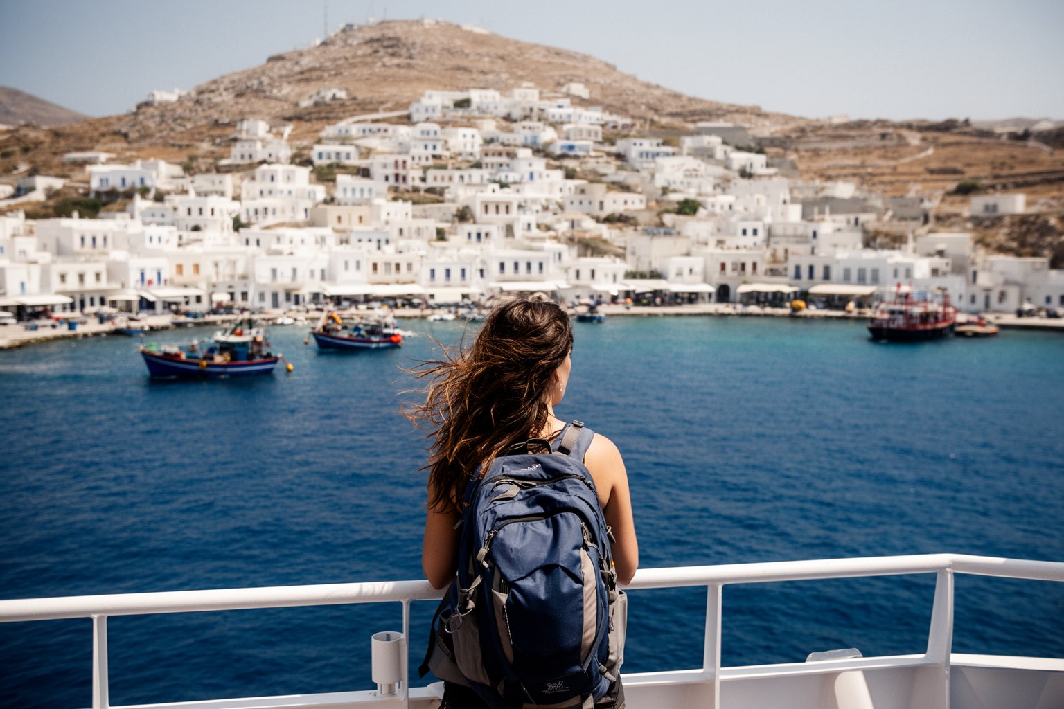Solo Traveling Pros and Cons: Perks, Pitfalls and Tips Solo traveller with a backpack on a ferry deck as it approaches a whitewashed Greek island harbour